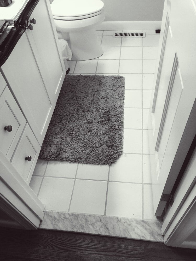 White tiled bathroom floor with dark countertop and plush bath mat in a home remodel by At Design Remodel in Mashpee