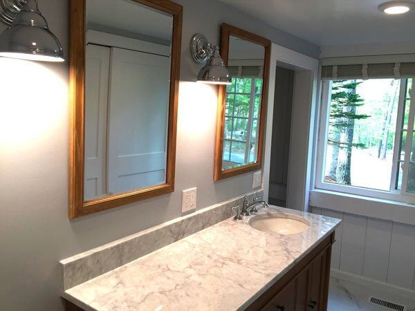 Completed bathroom remodel in New Seabury with marble countertop, wooden-framed mirrors, and a large window by At Design Remodel