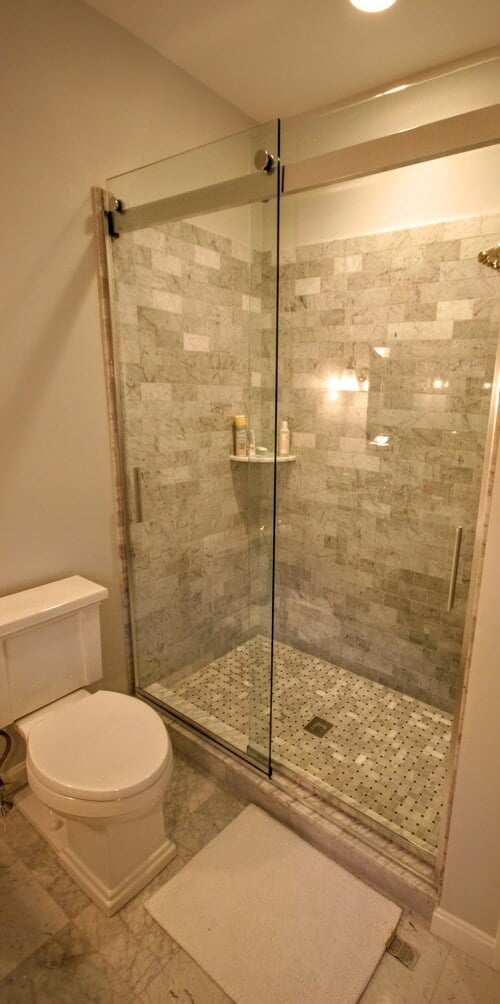 Marble-tiled shower with glass enclosure and rainfall showerhead in New Seabury Mews condo, remodeled by At Design Remodel