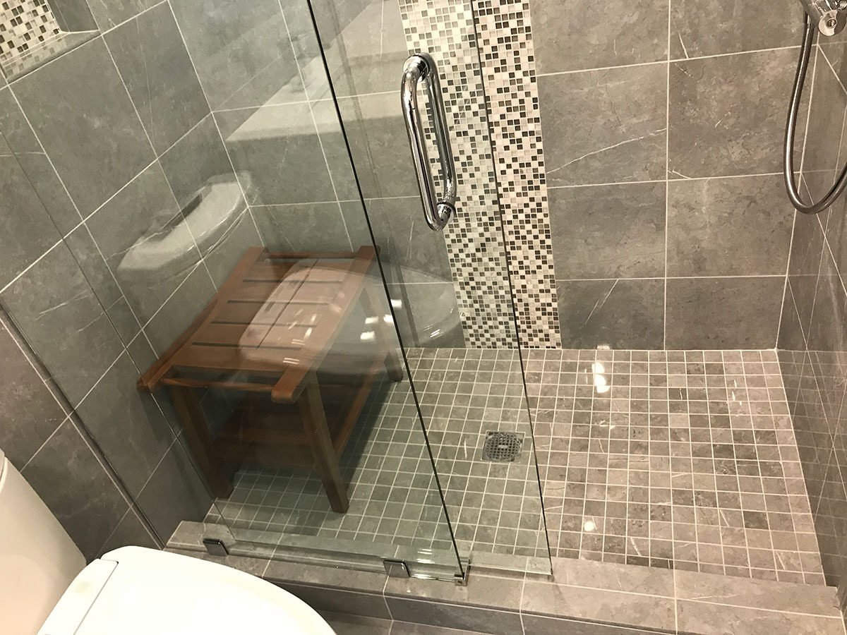 Gray tiled shower featuring glass doors and a wooden bench in a Yarmouthport bathroom renovation by At Design Remodel